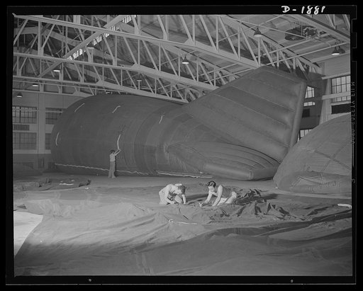 Barrage balloon manufacture. Final patching and tying. Men in the foreground are rolling cemented patches into correct position on the outside walls of a barrage balloon, while their co-worker in the background is checking the wrapping of ropes in cloth to prevent any stray bristles from puncturing 