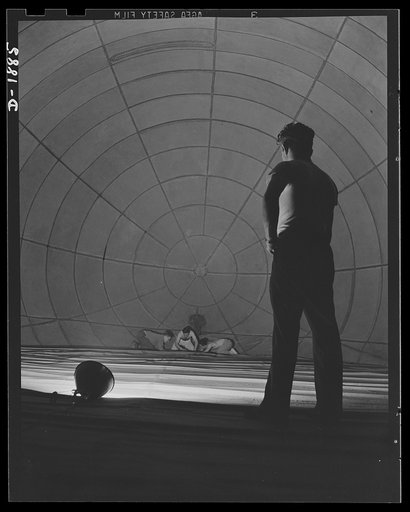 Barrage balloon manufacture. John and his colleagues. Silhouetted against the spiderweb pattern of the inside of a great barrage balloon, this supervisor watches "balloon tailors" at work on an inside seam. The area in the picture will be filled with helium in service. General Tire and Rubber Compan