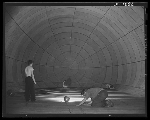 Barrage balloon manufacture. Spider and the flies. Putting last touches on a completed barrage balloon are these workers and their supervisor (standing). To inspect the bag for leaks, the light inside is turned off, leaving the workers in a midnight gloom. Then a co-worker on the outside takes a bri