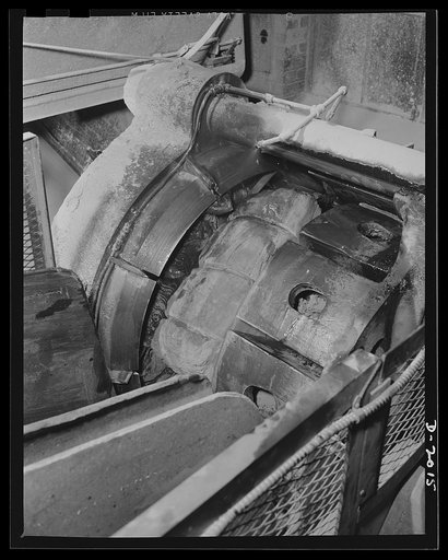 The guillotine. Having slid down the trough on the lower left, the bale  of raw rubber is now being squeezed against the knife blades on the left under terrific pressure. These blades fan out into eight selections, cut the rubber into eights like an orange divides into sections. General Tires (Fires