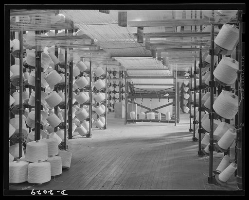 View of the creel room. Spools of cord are brought from storage into this creel room, from which they are woven into tire fabric. Each spool is connected by a complicated pulley arrangement to the bank of cords seen in the center of the picture and travels overhead to reach that bank. Firestone (Gen