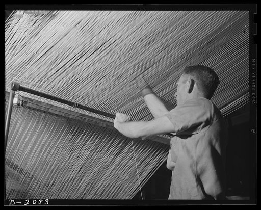 Splicing the line. The loose end in the worker's left hand is feeding an uninterrupted supply of cord to the machine from an emergency spool at the worker's feet. Meantime he searches through the maze of cords to find the one which has broken, to make his splice, and to resume the supply from the re