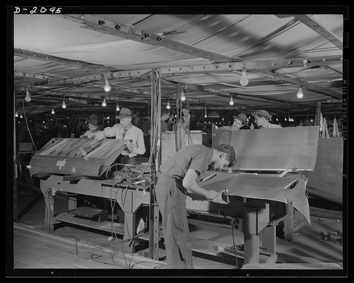 Subcontracting aircraft parts. These are aluminum flaps for military aircraft. The workers are in one of the canvas covered shops created in a former airship dock. The shops are enclosed to facilitate heating, since it would be impossible adequately to heat the entire air volume of the building, whi