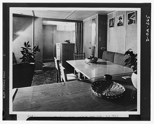 An interior of the Tennessee Valley Authority (TVA) experimental trailer-house, looking through the living-dining room into the kitchen. Walls, ceilings and floors are plywood, and the floor coverings are linoleum. Of the numerous built-in items of furniture and equipment in the house, the picture s