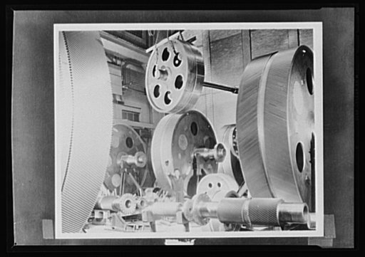 Gears to grind the Axis. Giant gears to guide America's fleets around the sees to victory. A workman, dwarfed by the towering wheels, directs the moving of a high speed gear blank to a hooking machine. The surge of the mightiest surf won't faze these titans, whose job it is to speed the day when the