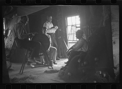 Untitled photo, possibly related to: Fishermen in repair shack repairing nets and sharpening tools. The twine of these nets is impregnated with coal tar; as the sewing of nets requires the holding of the twine in the mouth an occupational disease has developed which is referred to as fishermen's lip