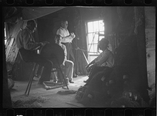 Untitled photo, possibly related to: Fishermen in repair shack repairing nets and sharpening tools. The twine of these nets is impregnated with coal tar; as the sewing of nets requires the holding of the twine in the mouth an occupational disease has developed which is referred to as fishermen's lip
