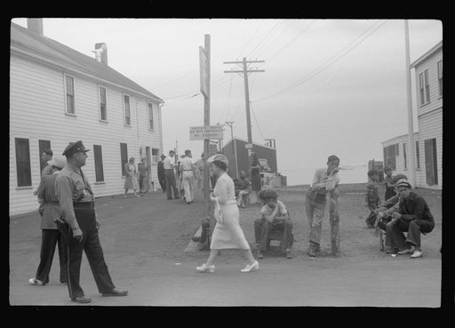 Untitled photo, possibly related to: Entrance to the town pier before the arrival of the Boston boat. The policeman is an institution, knows everybody, even every summer visitor. The boys wait for the chance of carrying somebody's baggage. Provincetown, Massachusetts