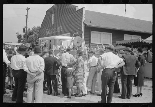 Groups of people standing in front of weighing concession with owner trying to drum up trade, state fair, Donaldsonville, Louisiana