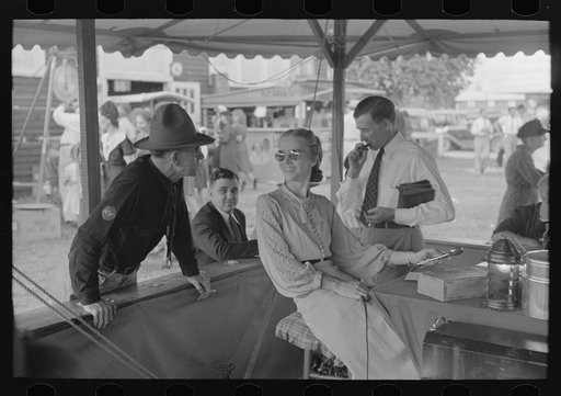In the game of bingo, an announcement is made by microphone as to the next number coming up. Microphone operator is shown here talking with an old friend who has just arrived. State fair, Donaldsonville, Louisiana