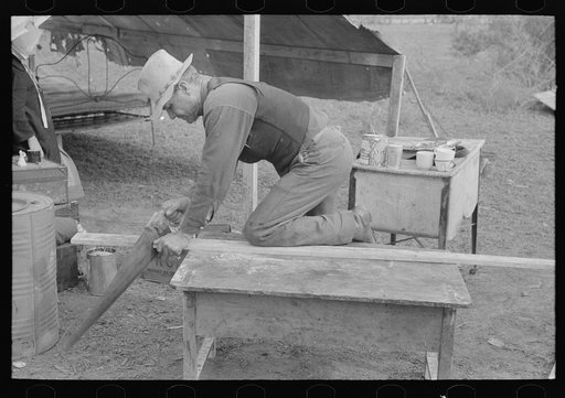White migrant worker sawing wood for stakes to be used in setting up tent home, near Harlingen, Texas