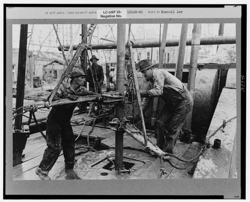 Oil roughnecks screwing one piece of drill pipe to another piece by means of a heavy pipe wrench, Kilgore, Texas