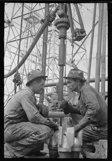 Oil drillers talking with bits in front of them and drilling equipment in background, Kilgore, Texas
