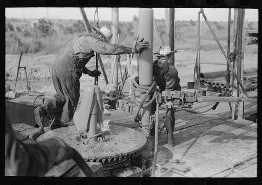 Adding a length of drilling pipe at oil well in the Seminole oil field, Oklahoma. Clamps are put in place; wrench applied to loosen pipe; mud spurting out as the sections of pipe are loosened; freed is moved over to rat hole; lower end of traveling block is attached to pipe in rat hole by means of r
