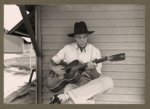Migratory worker playing guitar on front porch of his metal shelter in the Agua Fria Labor Camp, Arizona