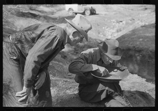 Gold prospector blowing away dirt to find the gold in his pan while a visiting prospector looks on. Pinos Altos, New Mexico