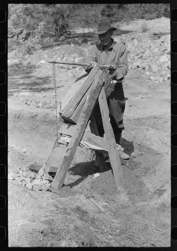 Prospector operating dry washer called a papago. This dry washer is the old Indian method of washing for gold. The rocks and dirt are placed in the higher compartment. Below and to the back is a bellows made of canvas. This is pumped to blow through the screening and thus blow away loose dirt while 