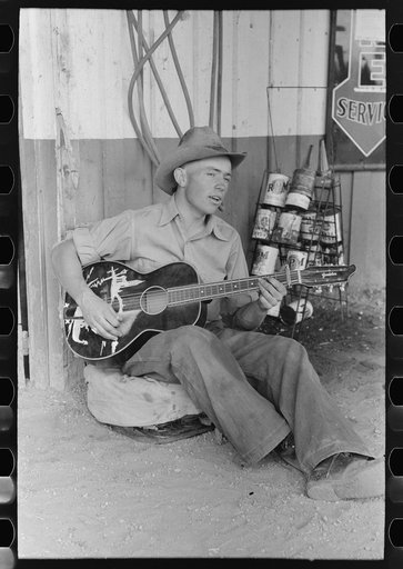 Farm boy playing guitar in front of the filling station and garage, Pie Town, New Mexico