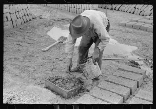 Adding a little water to adobe mixture in wooden form in process of making adobe bricks, Chamisal, New Mexico