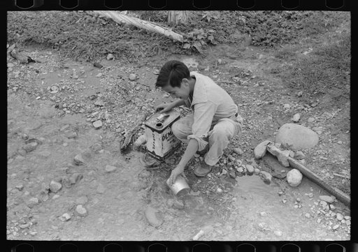 A Spanish-American boy dipping water from irrigating ditch. Water will be used in house for cooking, drinking, etc. Chamisal, New Mexico