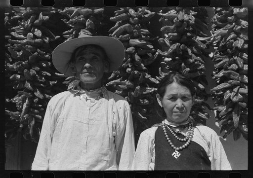 Indian and his wife in front of house with chili peppers, Isletta, New Mexico