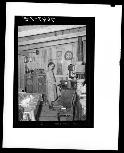 Mrs. Spiller at stove in their one-room shack. Note narrow window. Also Mrs. Spiller wearing overshoes because of dampness of dirt floor. Southeast Missouri