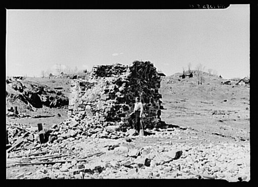 Mr. Henrickson, one of the old timers at Mansfield, Michigan, standing at the base of the former one hundred foot smokestack of an abandoned iron mine. This is one of the few recognizable remains of the mine
