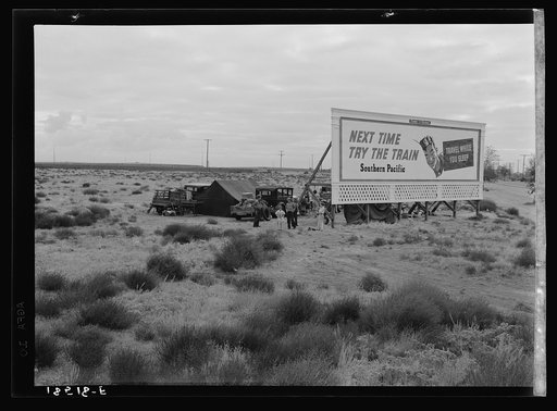 Three families camped on the plains along U.S. 99 in California. They are camped behind a billboard which serves as a partial windbreak. All are in need of work