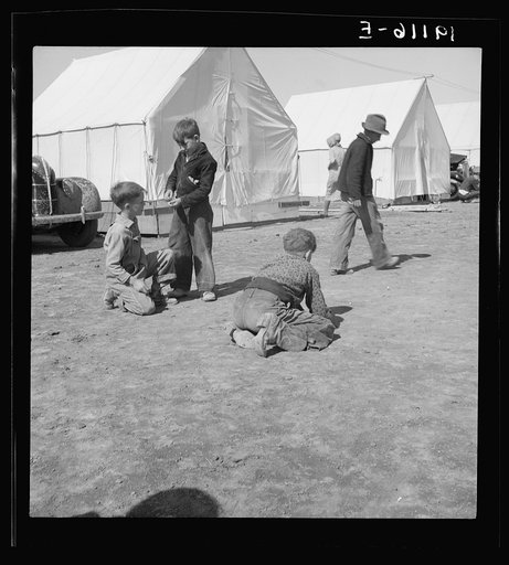 Migrant children in Farm Security Administration (FSA) migratory labor camp. Note type of boots worn by boy shooting marbles which tell his state of origin. Near Calipatria, Imperial County, California