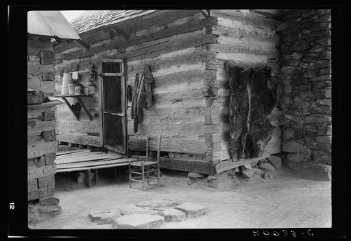 Construction detail of double log cabin of Negro share tenants. The cowhide was hung there after being dried on a barn to be used as floor covering. Shelf shows churn, also bucket of water in which baby's bottle is kept cool. Person County, North Carolina