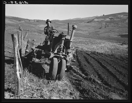 Young farmer plowing while other co-op members work in the sawmill. The tractor does work for five member families. Ola self-help sawmill co-op.  Gem County, Idaho
