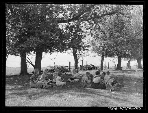 Teacher eats lunch with children in shade of trees on school grounds. Grundy County, Iowa