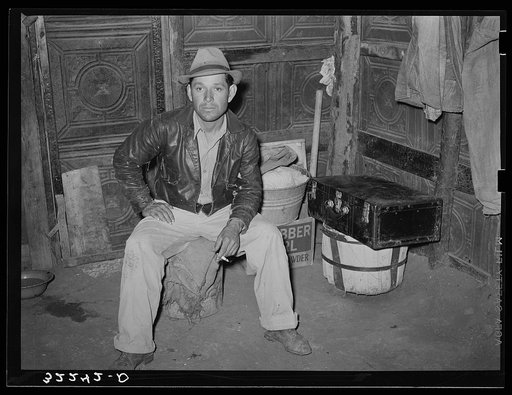 Mexican man in corner of one-room dwelling. Notice dirt floor and siding of house which is constructed of discarded tin ceiling material. Robstown, Texas