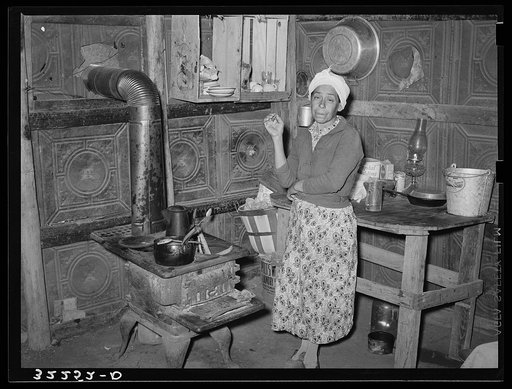 Mexican woman in kitchen corner of her one-room dwelling made out of discarded tin ceiling material. Notice dirt floor. Robstown, Texas