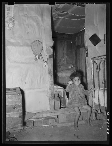 Mexican child sitting in doorway between living and bedroom in his home on the outskirts of San Antonio, Texas. Notice the dirt floor