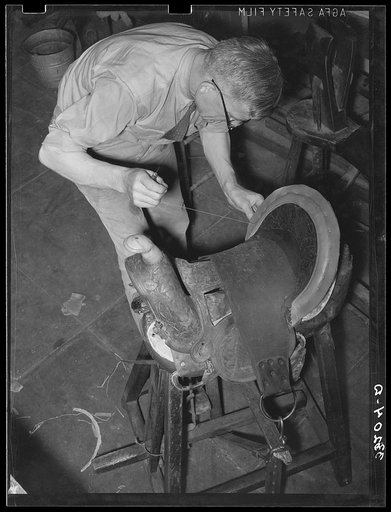 Saddle maker sewing in repairing saddle. Notice awl in his hand. Saddle shop, Alpine, Texas