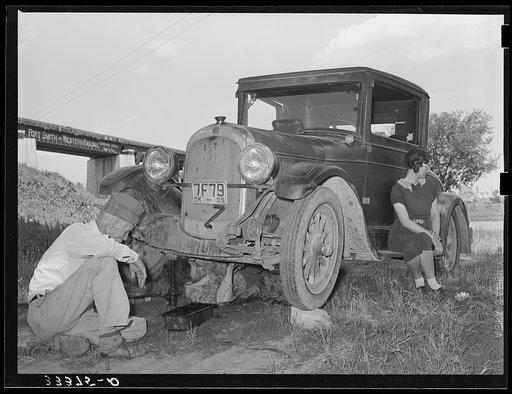 Migrant steeple-jack dropping the pan of his automobile before replacing burned-out bearing. His father and wife are also shown. When he secures a job, the entire family, consisting of his father and mother, wife and sister, all help with the painting. Camped near Prague, Oklahoma