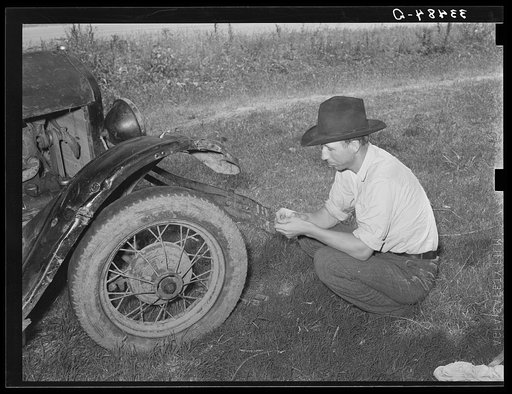 White migrant squatting down in front of his automobile while camped near Prague, Olkahoma. These people use the term "hunkering down" instead of "squatting." "Hunkering down" is a slang term from the Indians