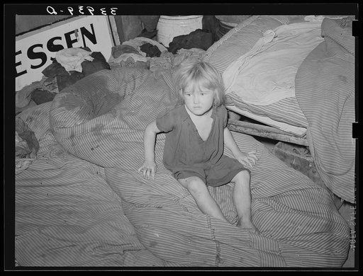 Child of large family sitting on bedding in shack home. Mays Avenue camp, Oklahoma City, Oklahoma