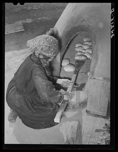 Spanish-American woman removing baked bread from outdoor earthen oven by means of a long wooden paddle. Near Taos, New Mexico