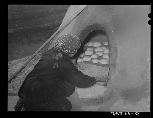 Spanish-American woman putting boards over entrance of outdoor oven to retain heat for baking bread. Near Taos, New Mexico
