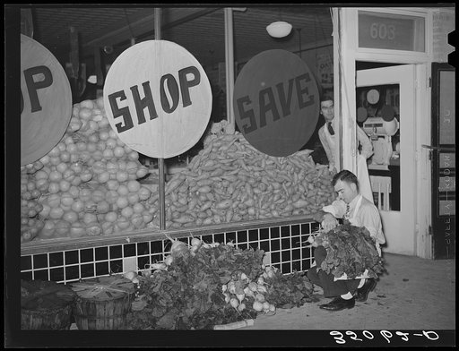 Trucks unload fresh vegetables from early morning market. The clerk takes them in from the sidewalks and arranges them on the display racks. Retail grocery, San Angelo, Texas