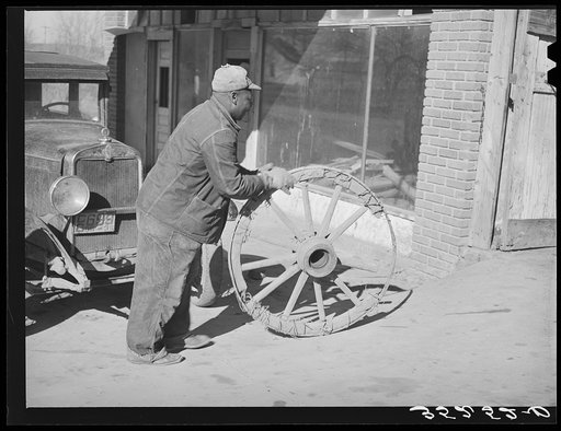 Negro farmer rolling much-repaired wagon wheel into blacksmith shop to see if it can again be repaired. Depew, Oklahoma