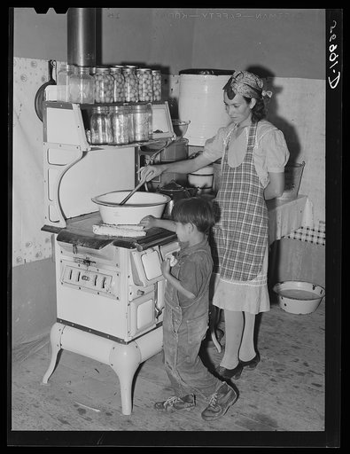 Spanish-American woman stirring pan of cooking beans. Small boy is roasting sweet corn on top of the stove. Chamisal, New Mexico