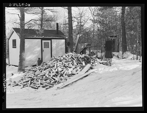 Untitled photo, possibly related to: Mrs. M. Lane, young wife of a shipyard worker, gathering some wood just outside her one-room shack. The shack and little privy were built for them by her father. Made of celetex board. Bath, Maine