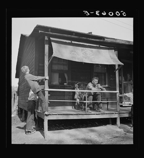 Former coal miner sitting on his front porch. He is the town philosopher, Jere, West Virginia. Repairs his home. Note railing and potato sack awning. Experiments with an elaborate garden on a hill about two miles away.  See 50081-E, 50057-E