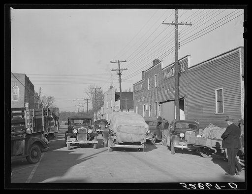 Street showing several tobacco warehouses on day of auction sales with farmers bringing in their tobacco on trailers and trucks. Mebane, Orange County, North Carolina