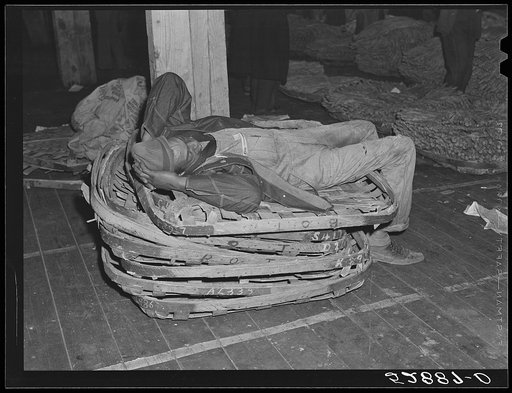 Worker in tobacco warehouse resting on top of empty tobacco baskets after auction sale. Durham, North Carolina