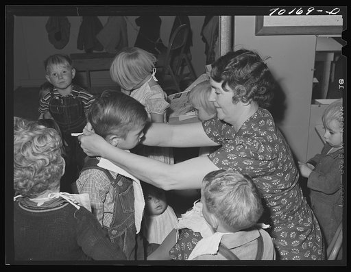 WPA (Work Projects Administration) teacher at the nursery school puts on bibs of her charges at the FSA (Farm Security Adminisrtation) family migratory labor camp. Yakima, Washington