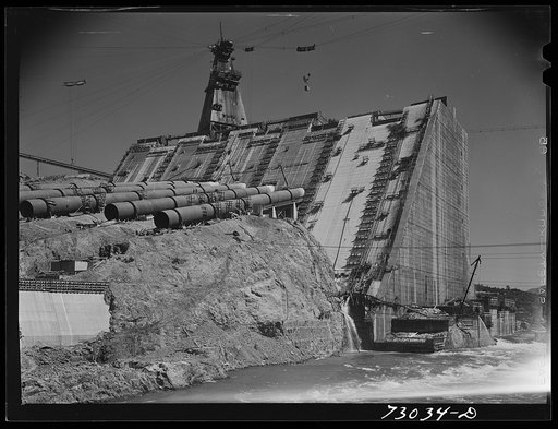 Untitled photo, possibly related to: Shasta Dam, Shasta County, California. One side of Shasta Dam under construction, with the Sacramento River still flowing through the opening between the two sides of the dam. Water will flow through the penstocks to the hydroelectric plant